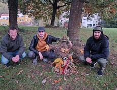 Drei junge Männer in herbstlich warmer Kleidung hocken neben einem selbstgemachten Naturkunstwerk, einem Männchen aus Stöcken und Blättern.