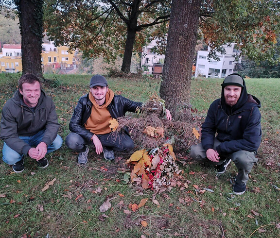 Drei junge Männer in herbstlich warmer Kleidung hocken neben einem selbstgemachten Naturkunstwerk, einem Männchen aus Stöcken und Blättern.