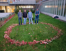 Vier Männer stehen auf einer Wiese auf der ein Mini-Fußballfeld mit Naturmaterialien aufgezeichnet ist und darum ist ein Kreis aus bunten Blättern.