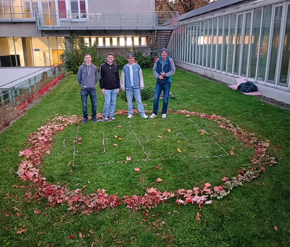 Vier Männer stehen auf einer Wiese auf der ein Mini-Fußballfeld mit Naturmaterialien aufgezeichnet ist und darum ist ein Kreis aus bunten Blättern.