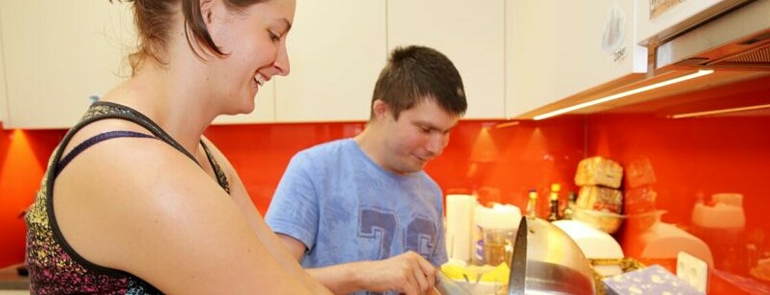 Eine junge Frau und ein junger beeinträchtigter Mann stehen in einer Küche und kochen gemeinsam.
